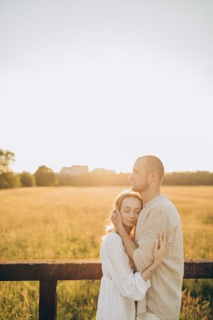 Tender embrace of couple in light colored clothing by wooden fence at golden hour.