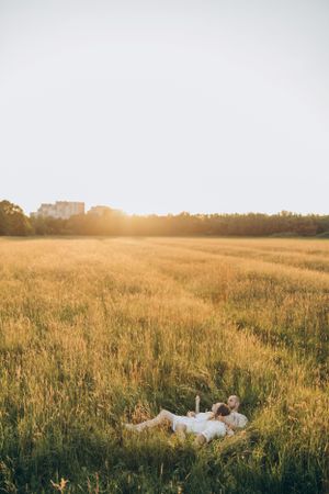 A couple lying in a golden wheat field during sunset with buildings in the distance.