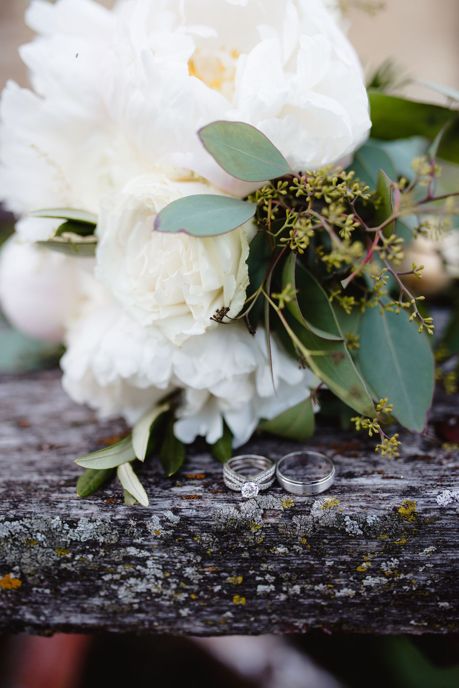 Wedding rings displayed with white roses and eucalyptus leaves on rustic wooden surface.