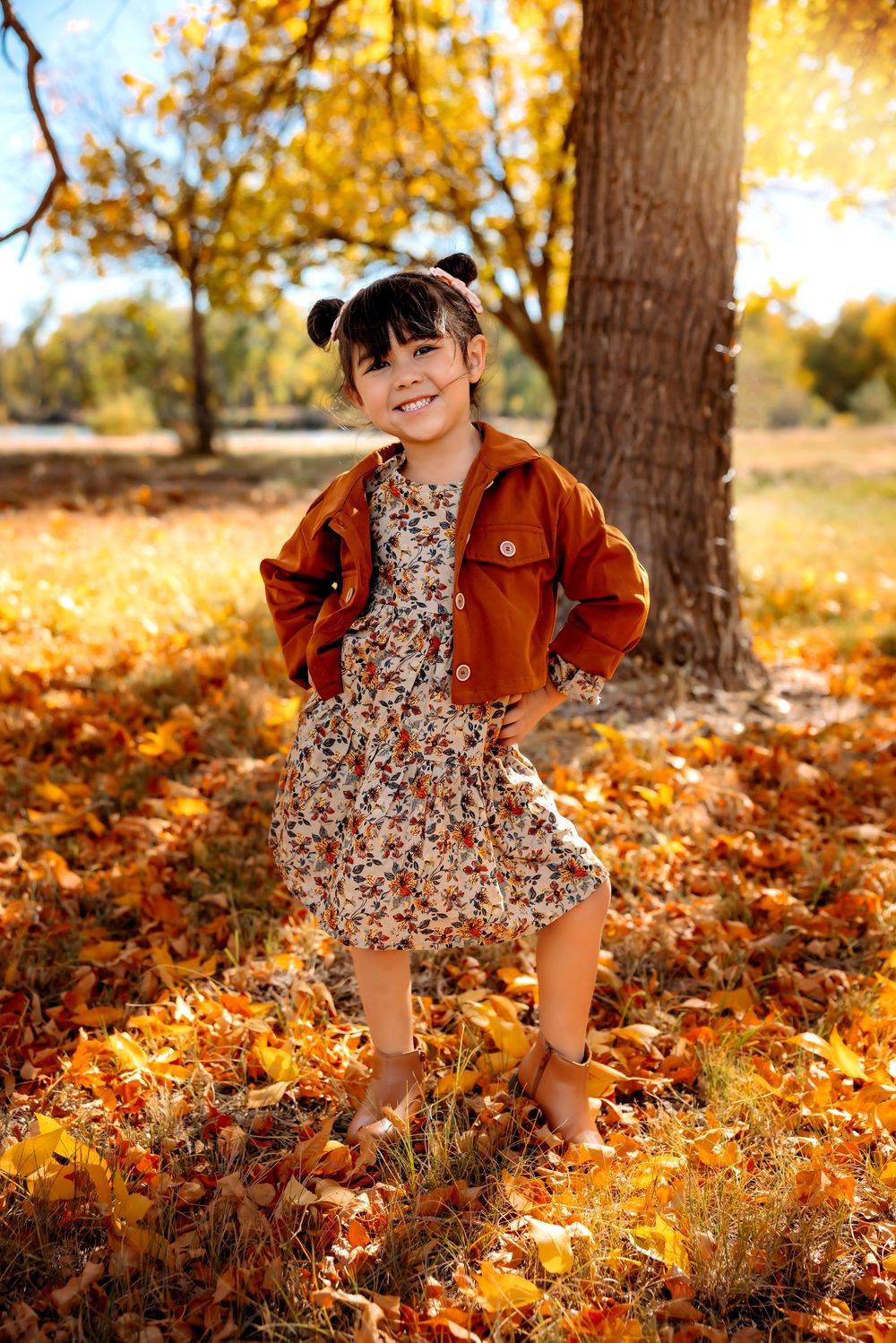 A child plays in a pile of golden autumn leaves beneath trees on a sunny fall day.