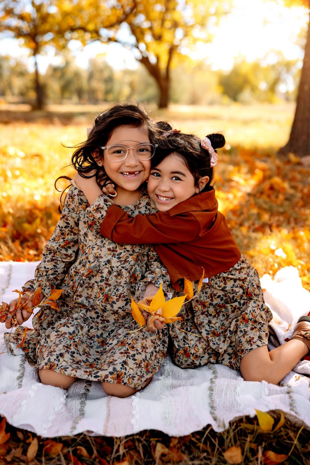 Two children in floral dresses sit on a white blanket surrounded by golden autumn leaves under a tree.