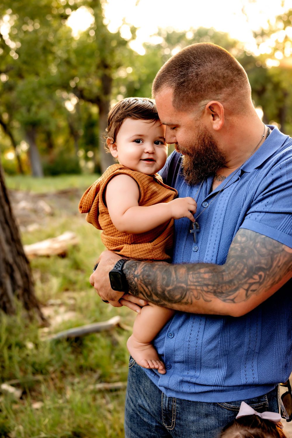 A parent in a blue denim shirt holds a small child wearing brown clothing in an outdoor setting during golden hour.