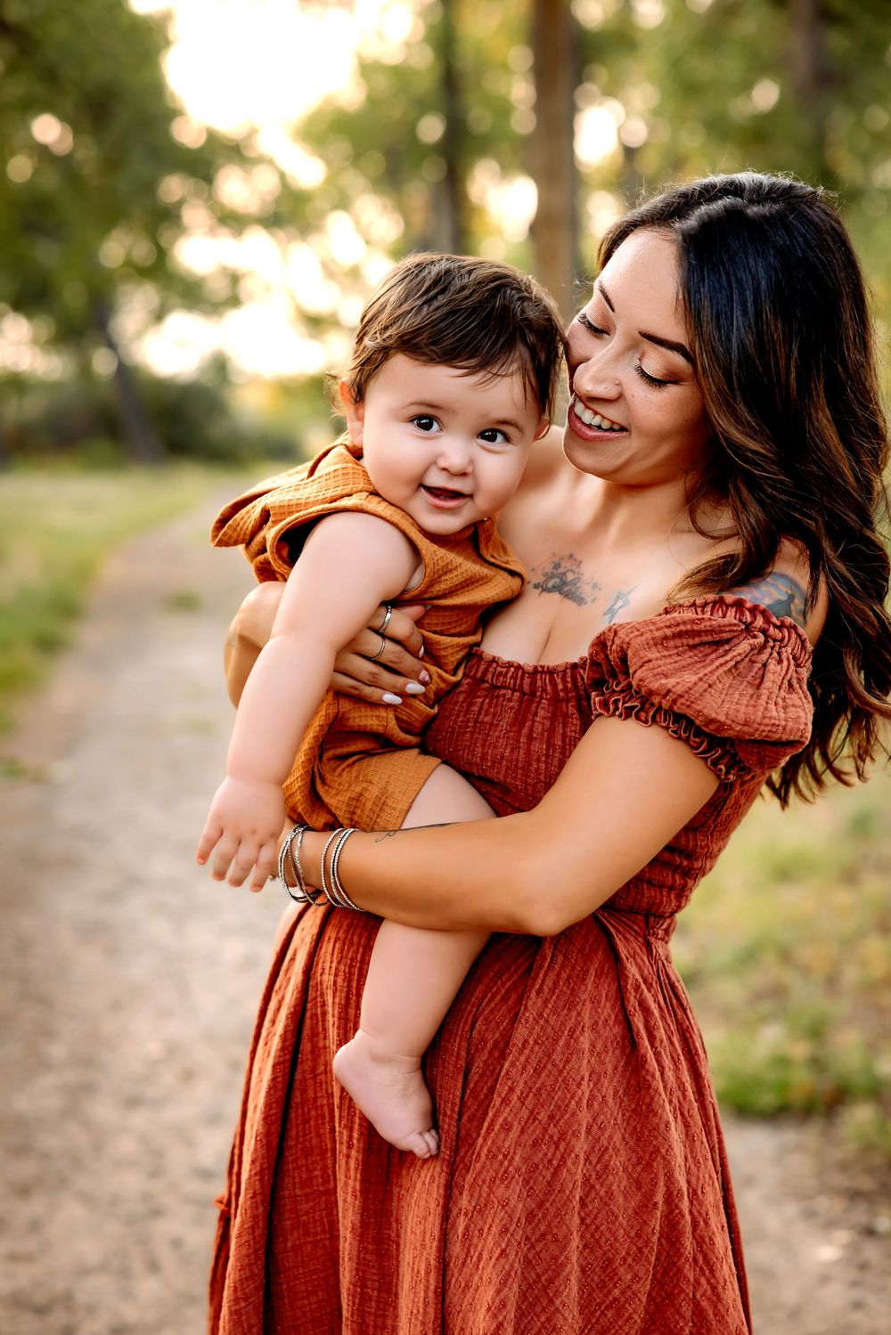 A tender moment captured on a nature path as a parent in a rust-colored dress holds a baby wearing brown at sunset.