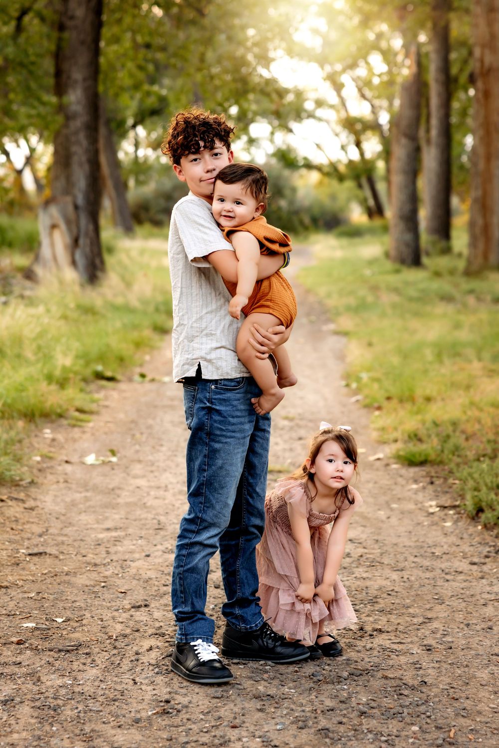 Three siblings pose together on a wooded trail during golden hour, with the youngest wearing orange and middle in pink.