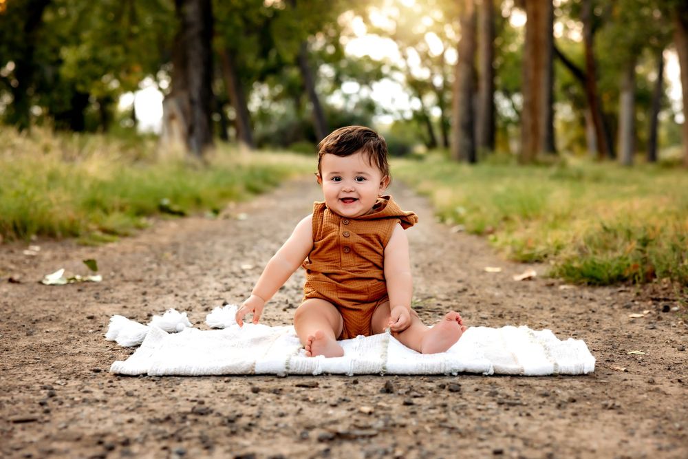 A baby sits on a white blanket outdoors surrounded by dirt and grass in a rustic setting.