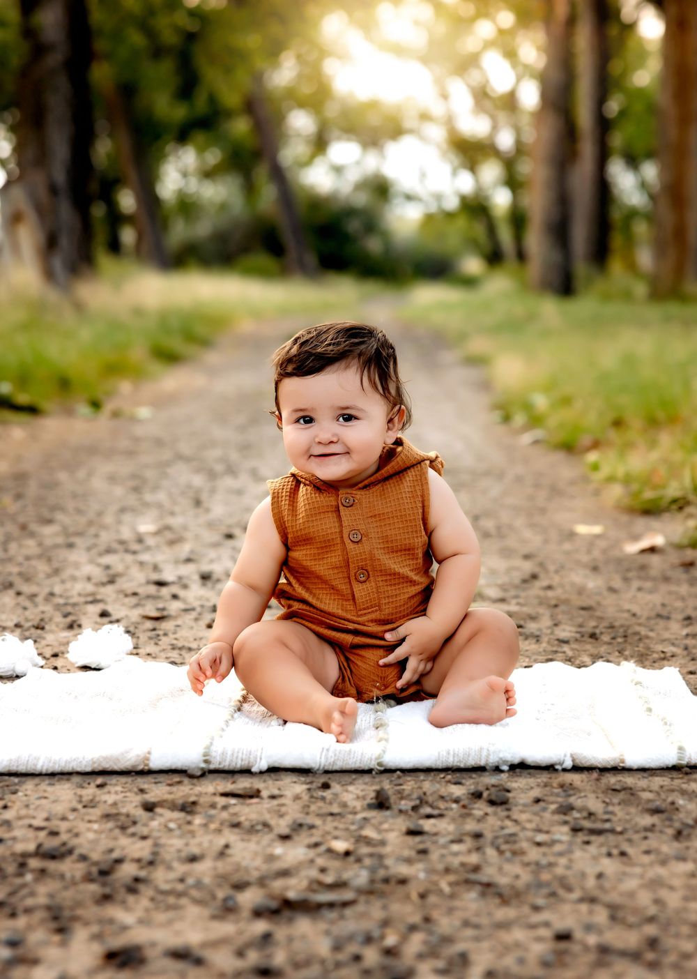 A baby in brown overalls sits on a dirt path with trees and bokeh lighting in the background.
