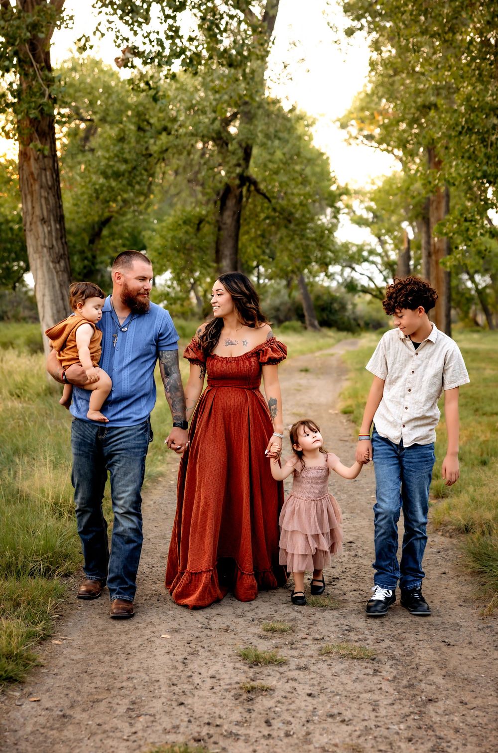 A family walks together holding hands along a dirt path through trees at sunset in matching casual outfits.