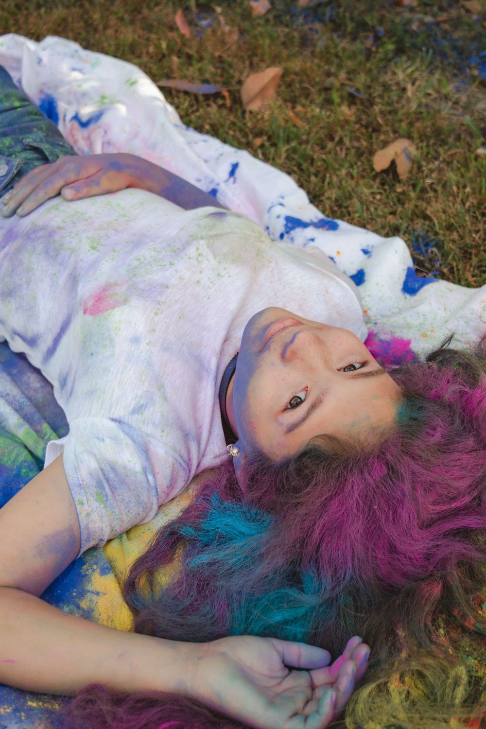 Person with colorful dyed hair lying on grass covered in paint powder.