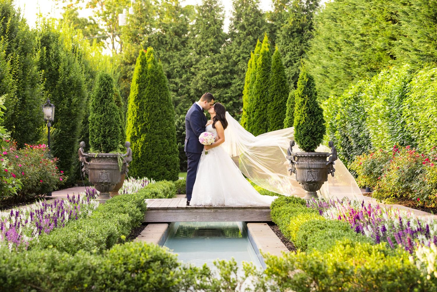 Garden wedding scene with formal pond surrounded by blooming flowers and cypress trees.