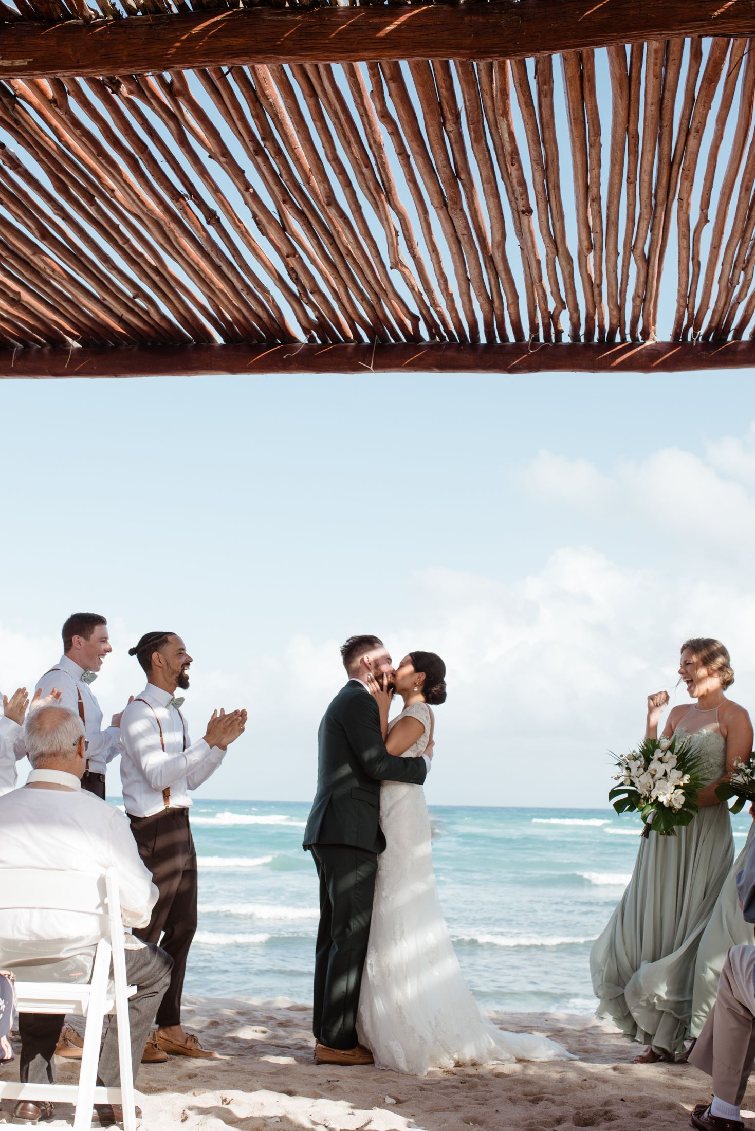 A romantic beachside wedding ceremony under a rustic palm frond canopy with ocean waves in the background.