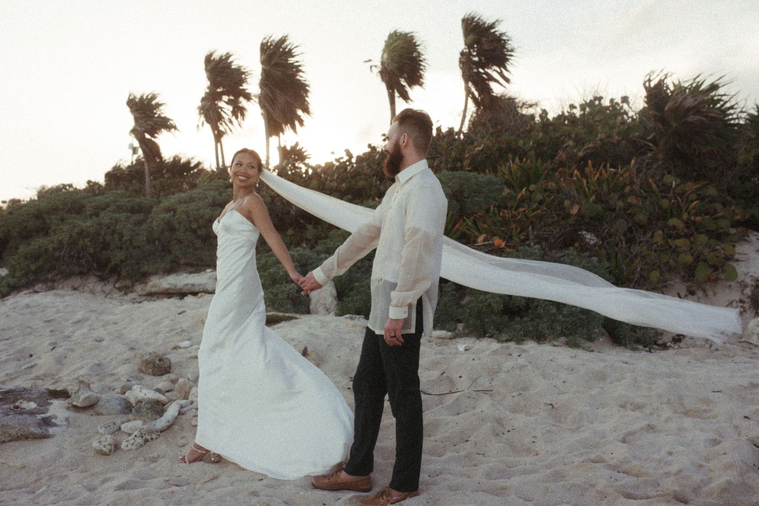 A couple shares a romantic moment on a beach at sunset with palm trees swaying in the wind.
