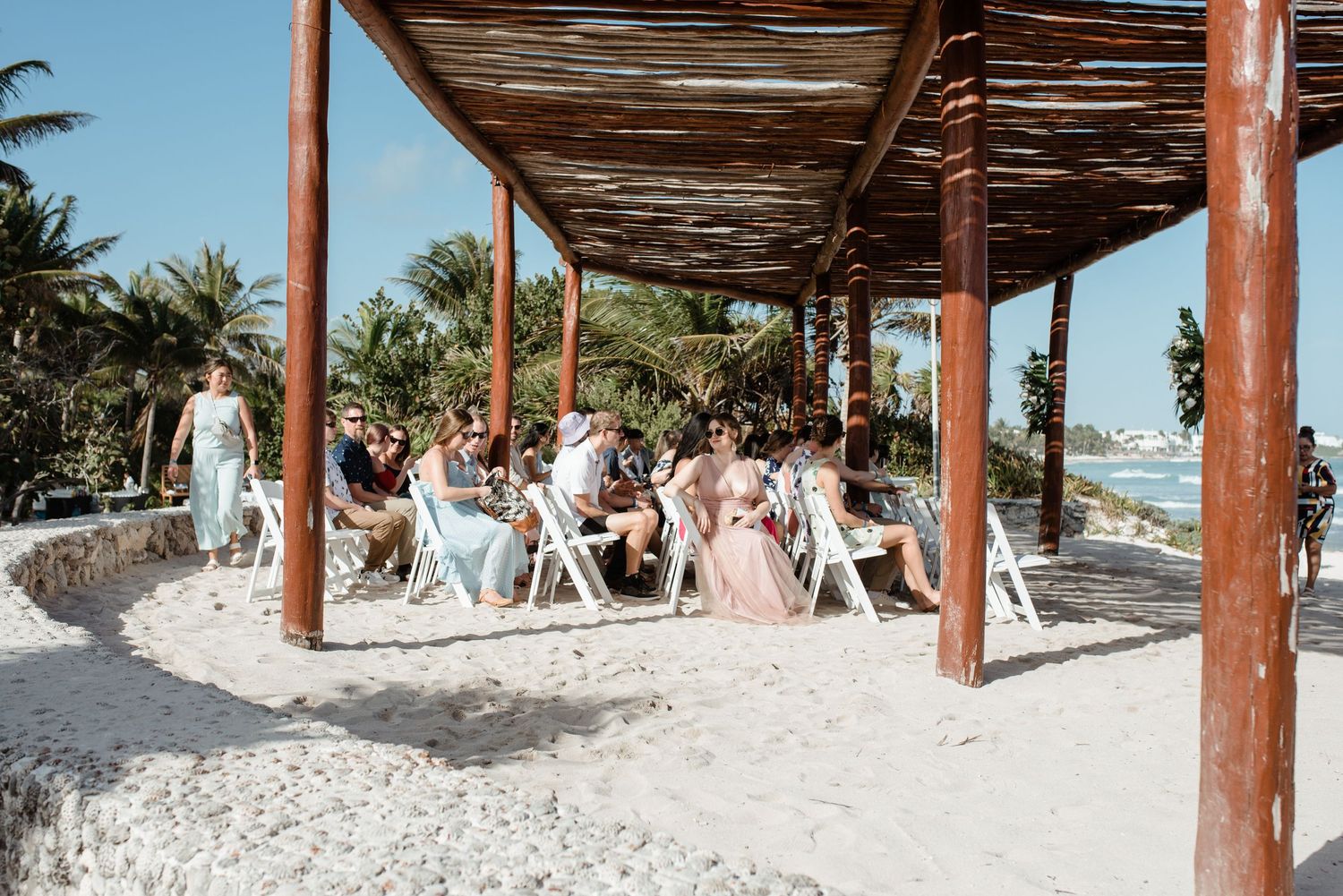 Beach wedding ceremony setup with wooden pergola and white chairs on sandy shore.