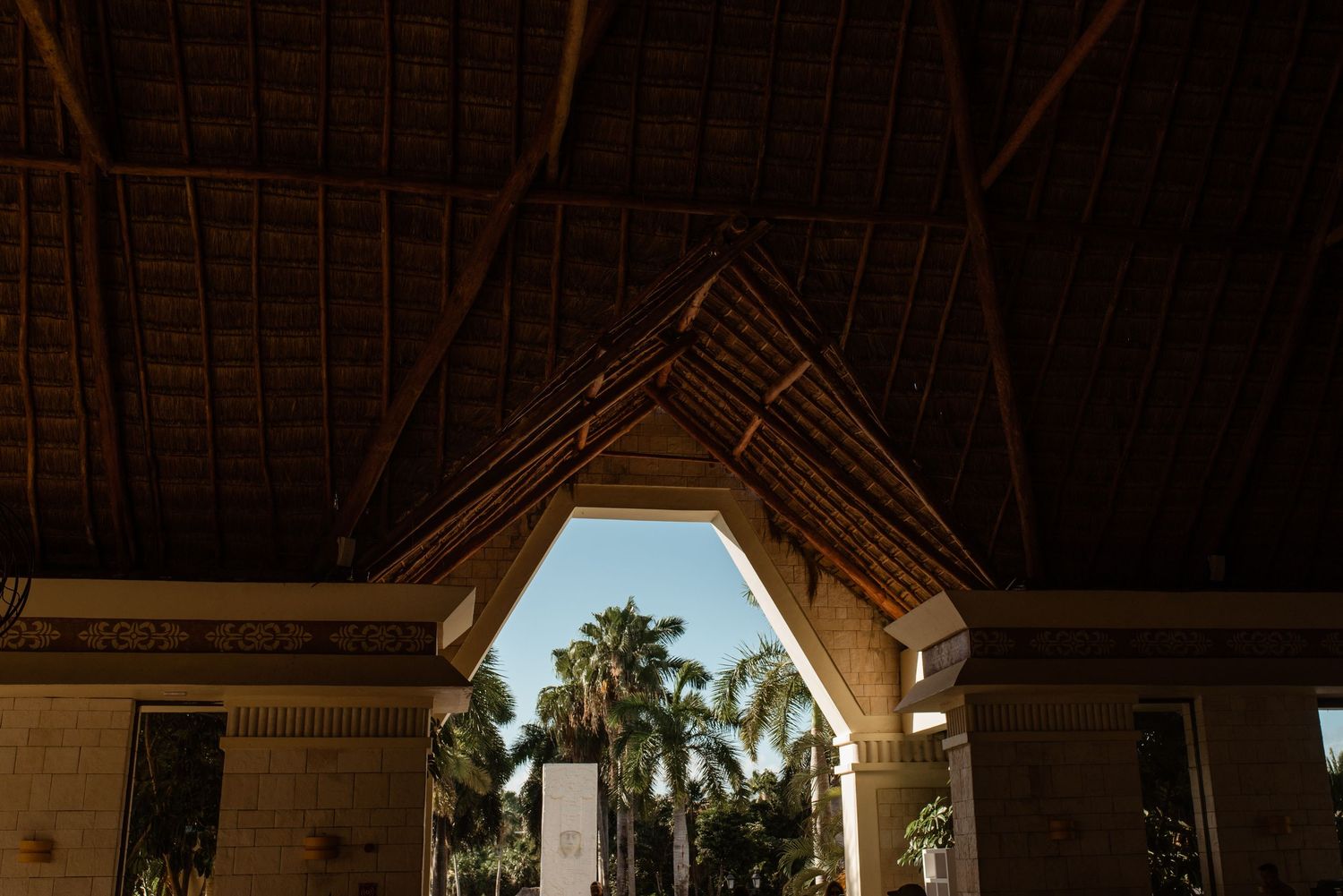 Architectural detail of a wooden thatched roof with palm trees visible through an arched opening.
