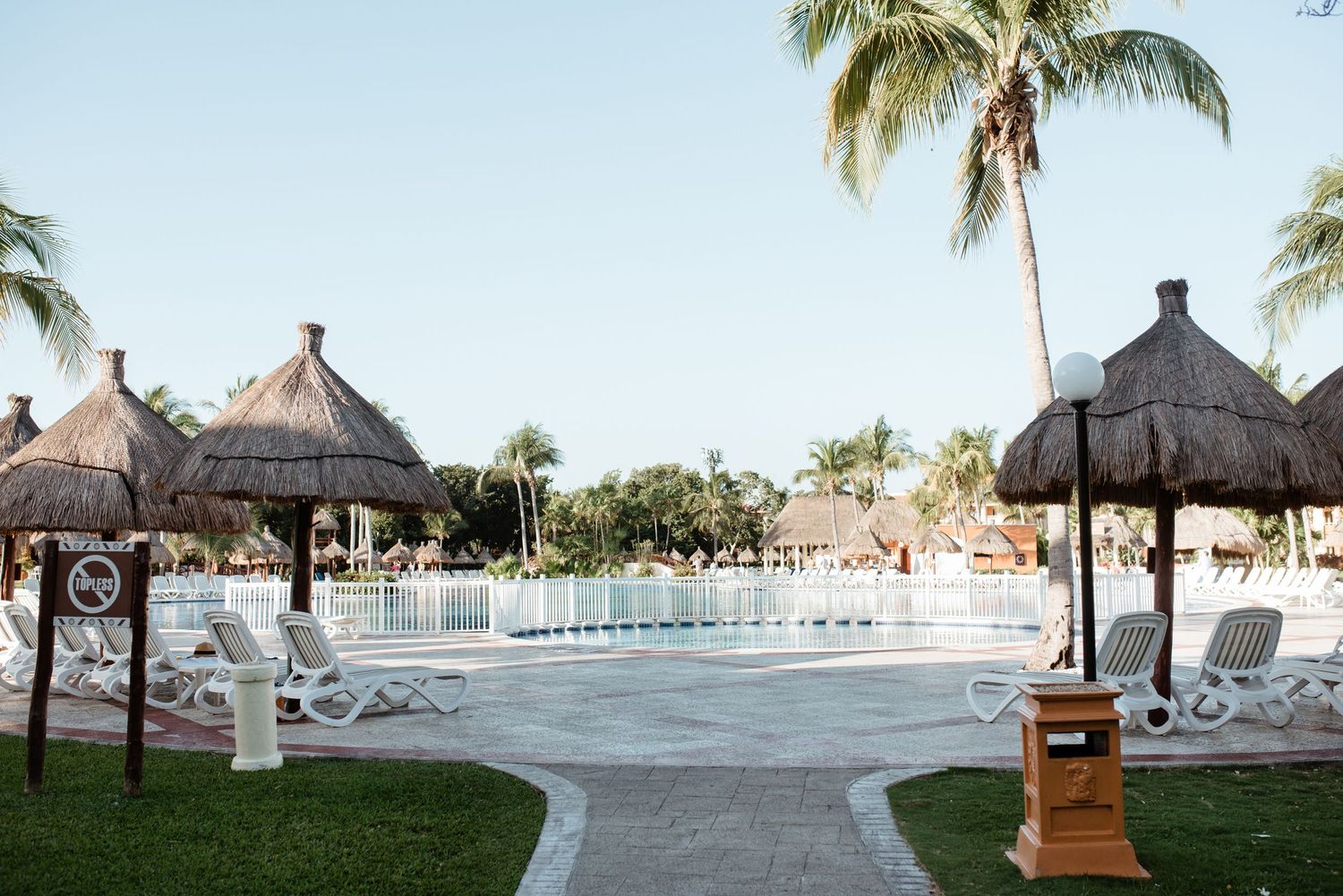 Resort pool area with thatched umbrellas and white lounge chairs arranged around a curved walkway.