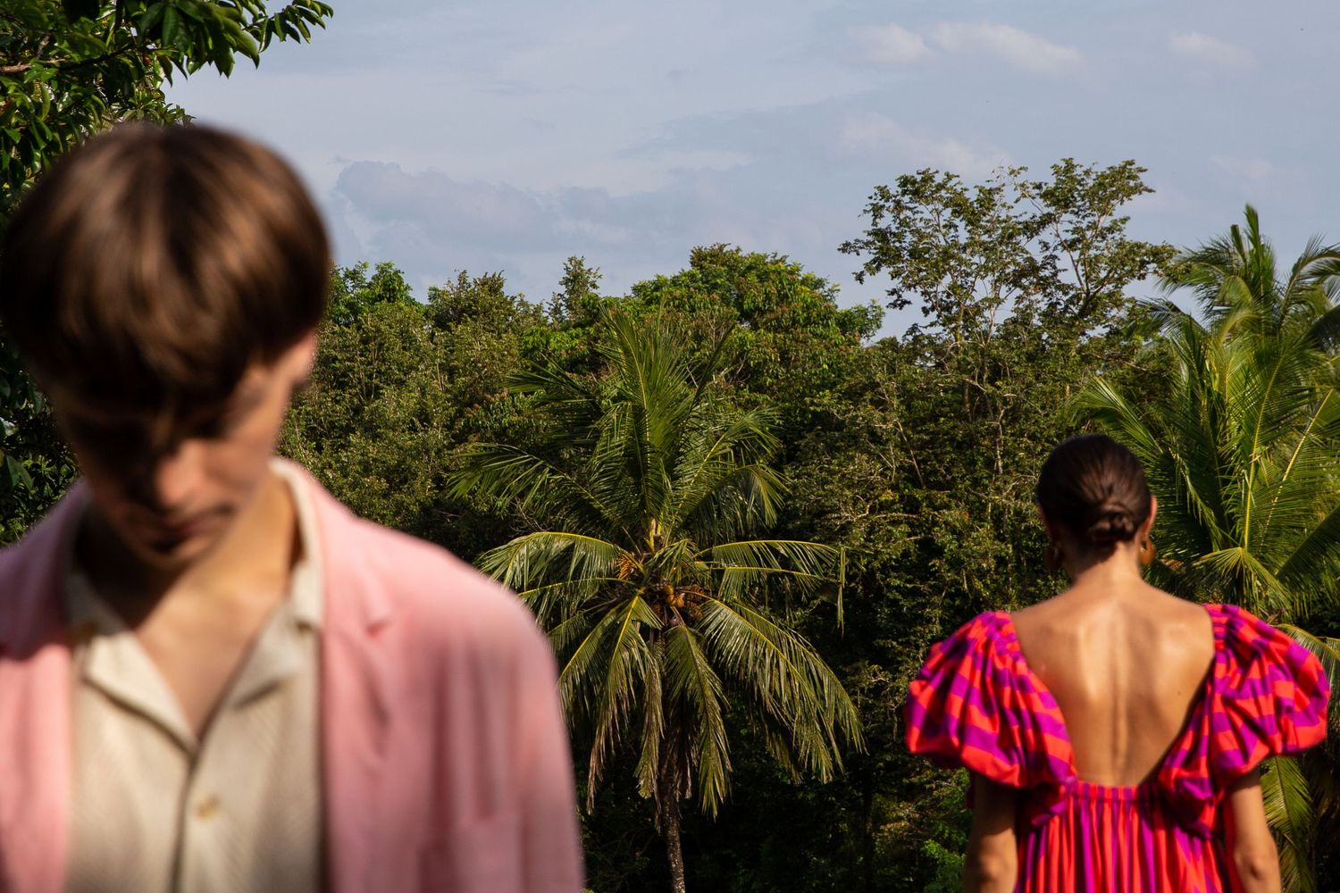 Two figures stand looking at a tropical landscape with palm trees against a bright sky.