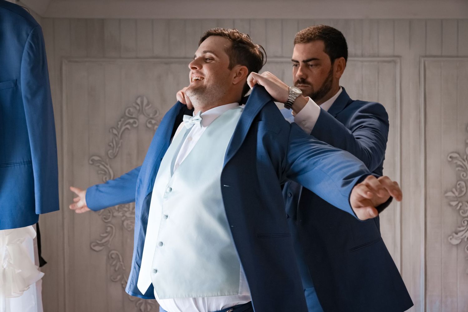Two men in blue formal wear getting ready in a white-walled dressing room.