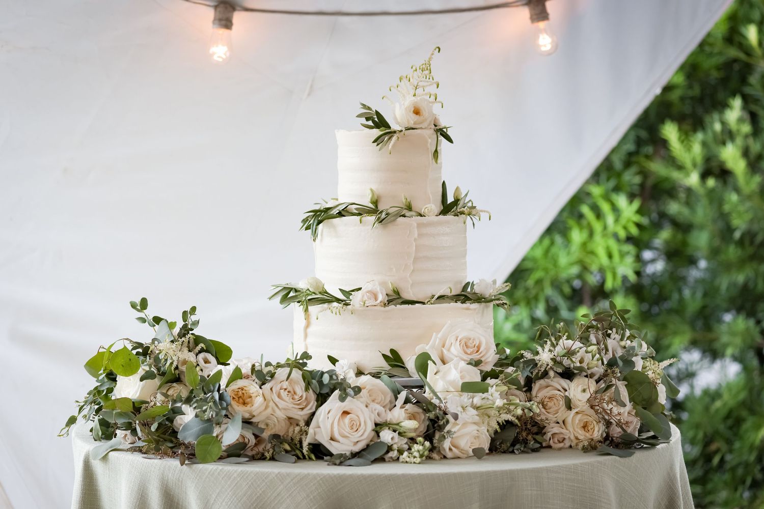 Elegant three-tier white wedding cake decorated with fresh flowers and greenery on white tablecloth.