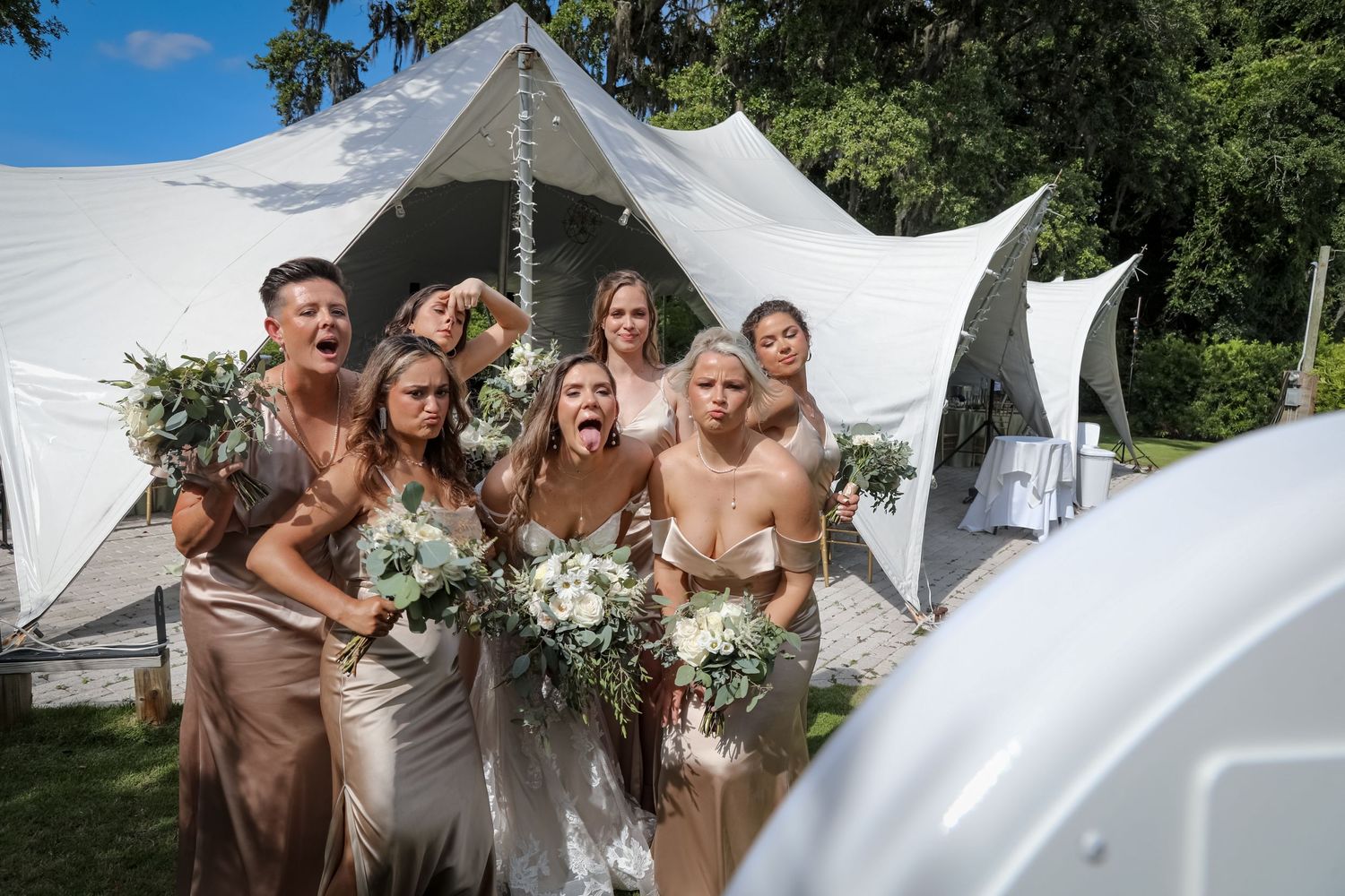 A wedding party poses together outdoors in front of elegant white tents on a sunny summer day.