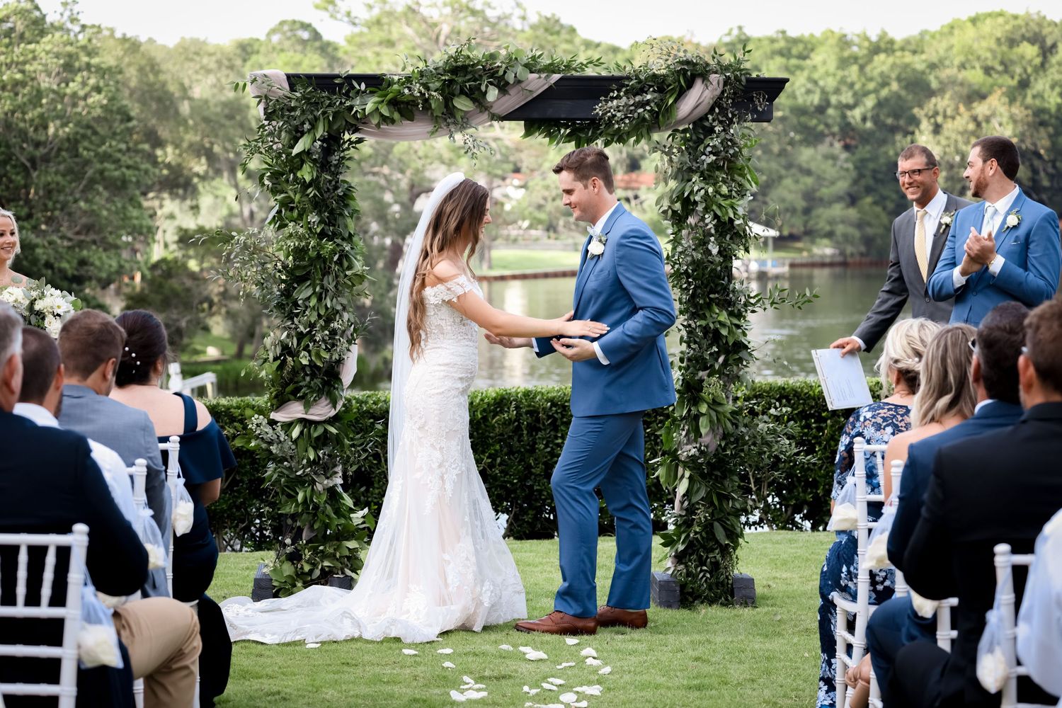 A romantic outdoor wedding ceremony takes place under a lush greenery arch beside a serene lake.