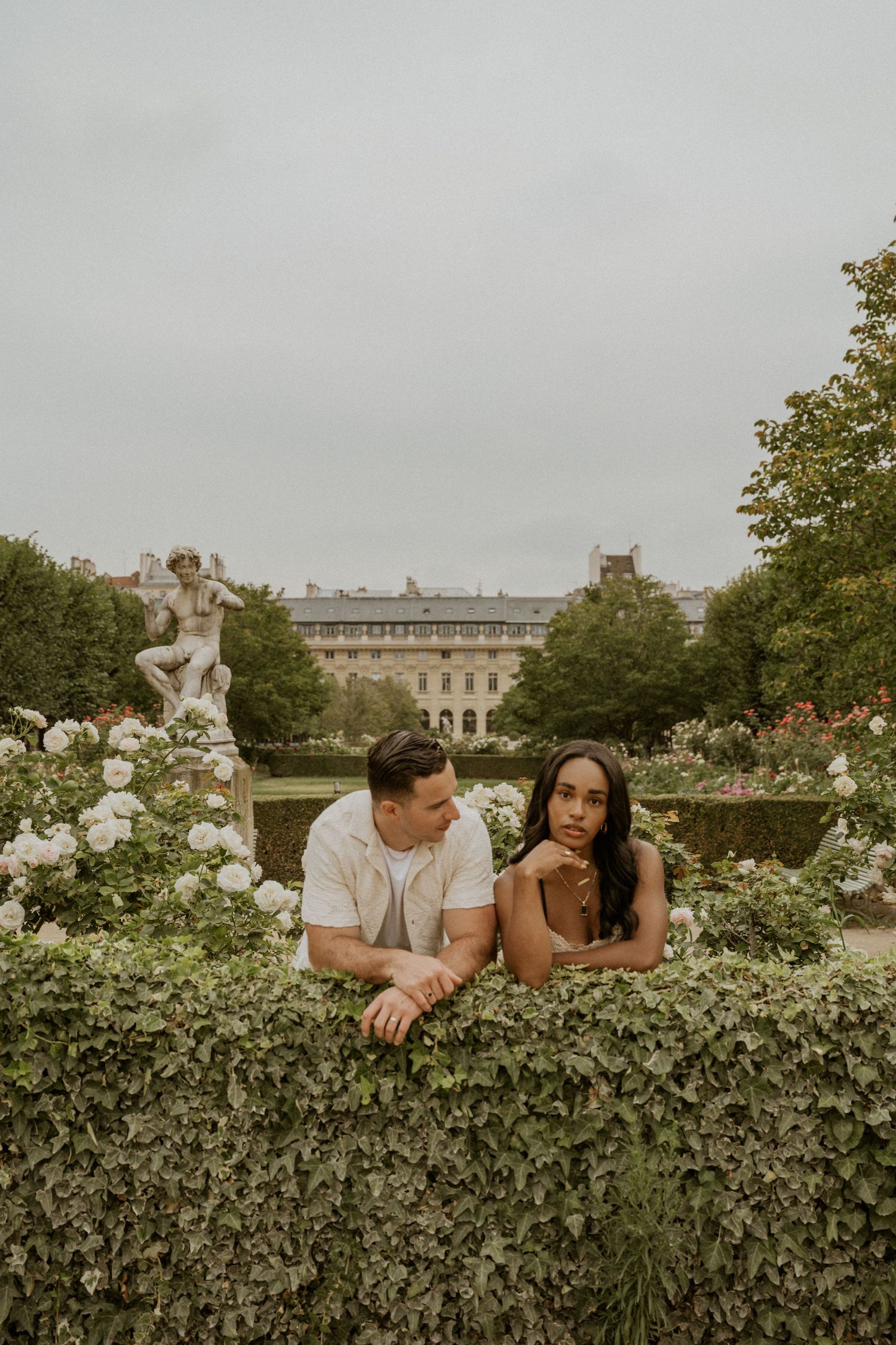 Romantic couple posing in garden with roses and historic building in background.
