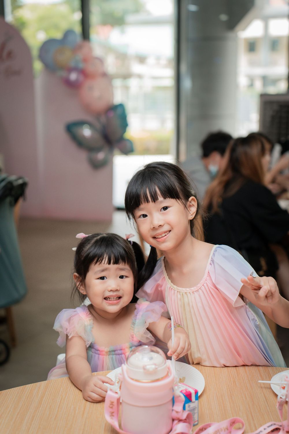 Two children in pink dresses share a special moment at a birthday celebration with a pink drink on the table.