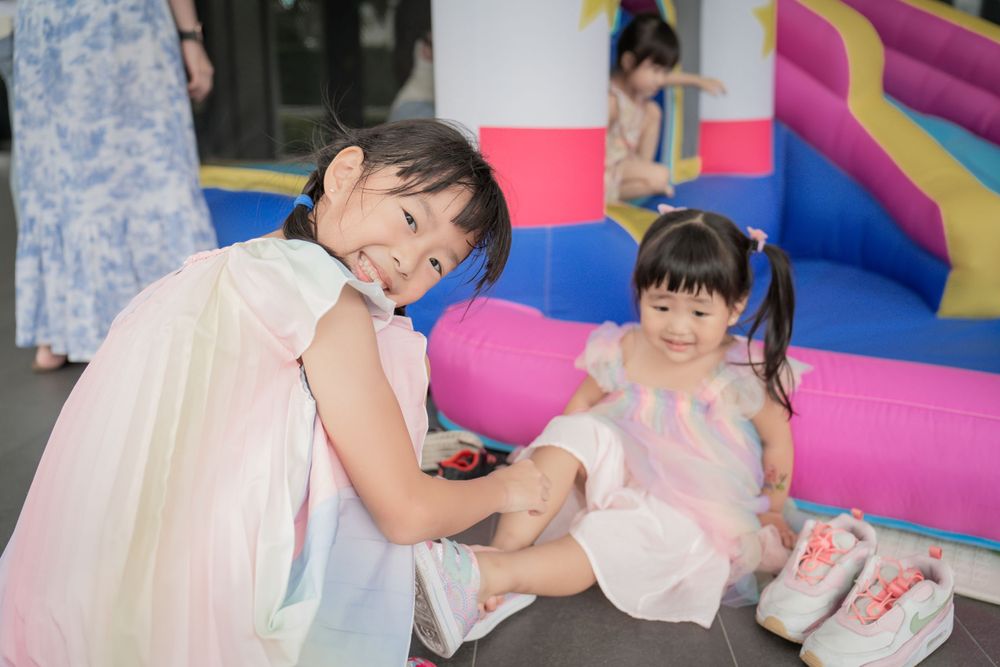 Two children in light dresses sitting and playing by a colorful inflatable bounce house.