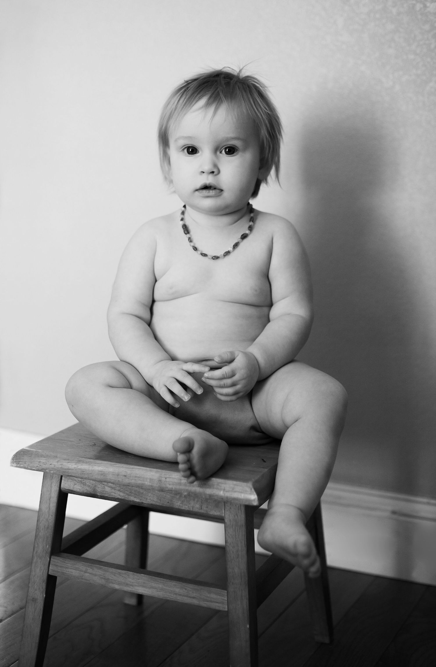 A black and white portrait of a toddler wearing a necklace while sitting on a wooden stool.