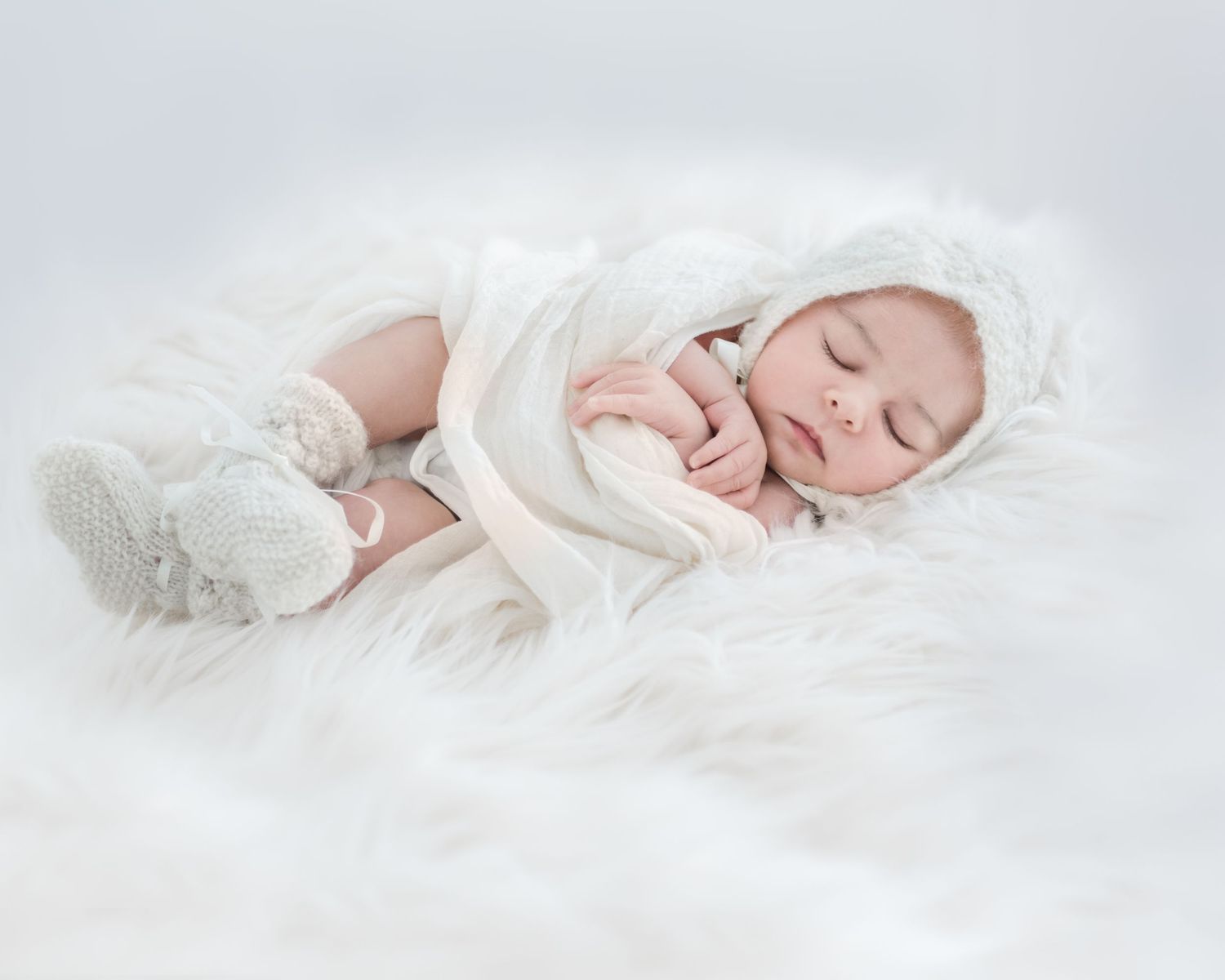 A sleeping newborn baby in white wrapped in soft blankets on white fur bedding.