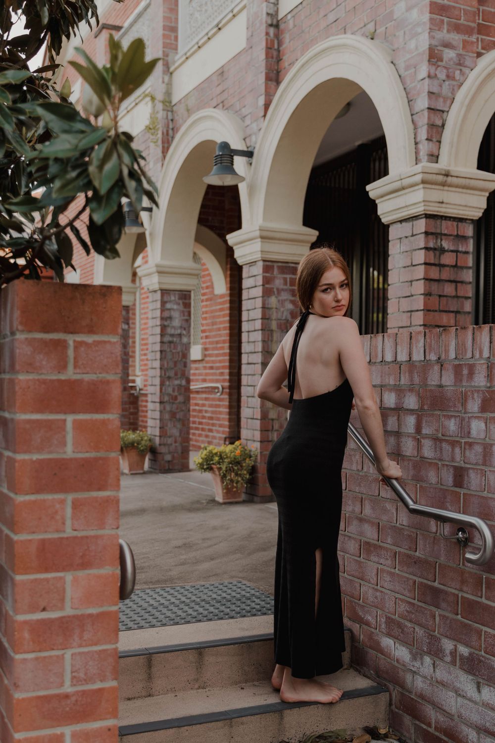 A fitted black evening dress posed elegantly on brick steps with arched architectural details.