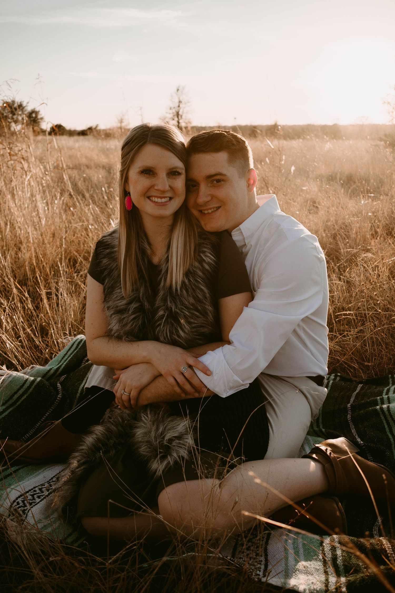 A couple sits together on a blanket in a field of tall grass during sunset.