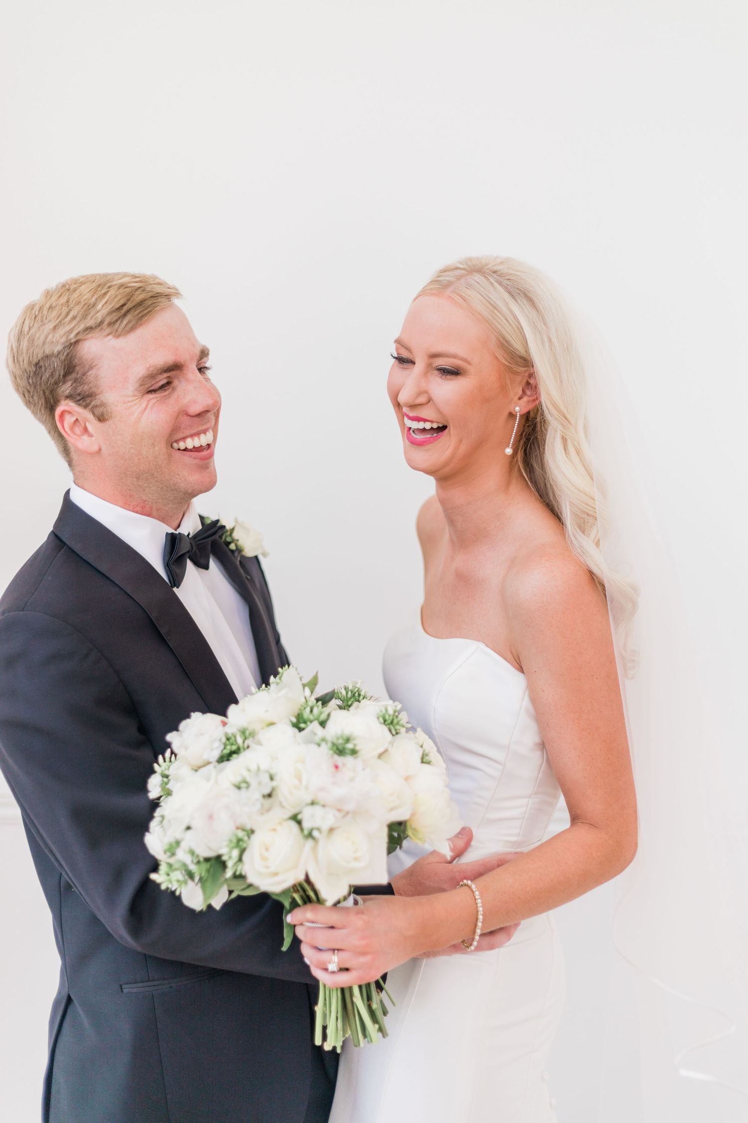 A newlywed couple sharing a joyful moment with a white bouquet against a light background.