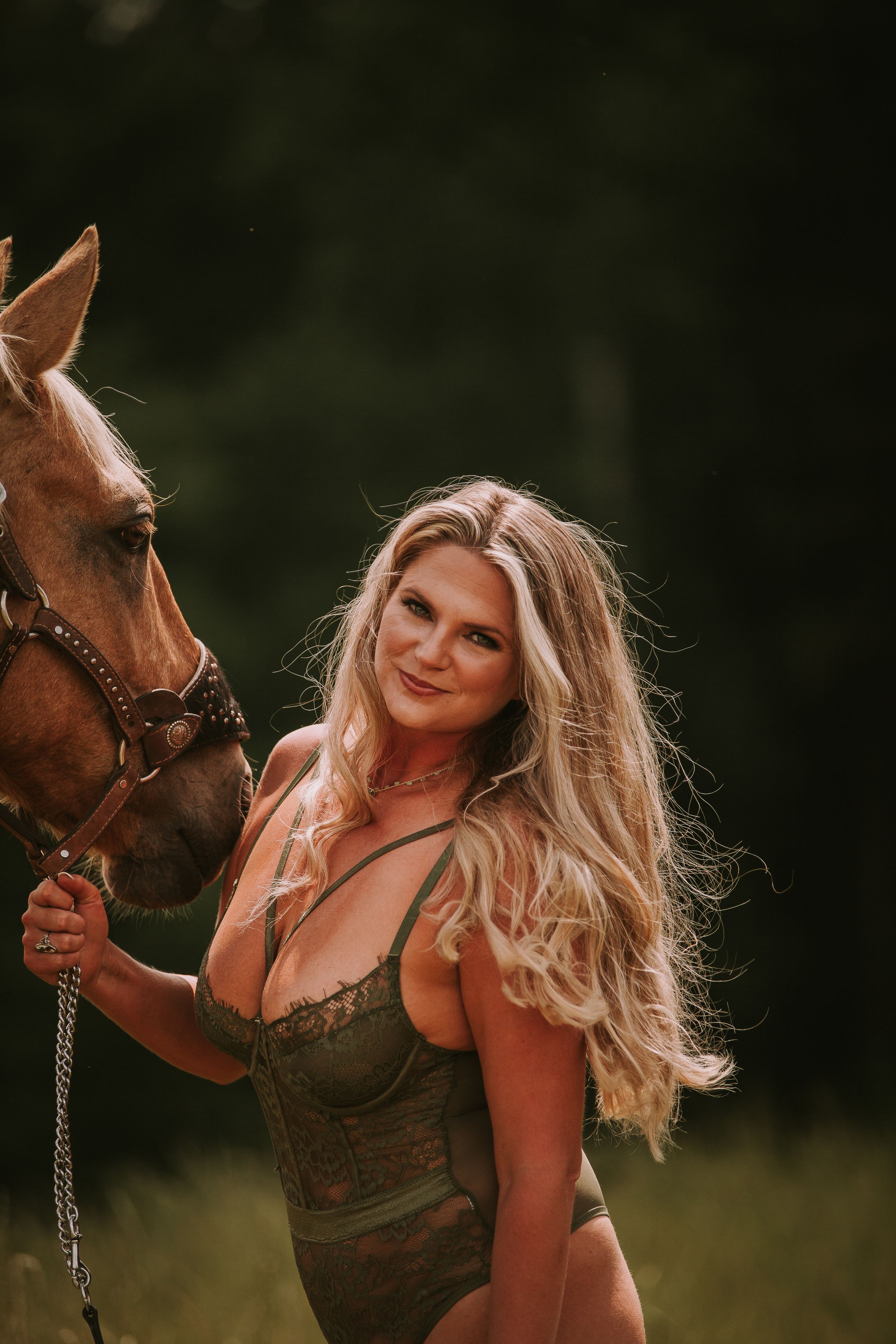 Woman in bodysuit poses with chestnut horse in rustic outdoor setting.