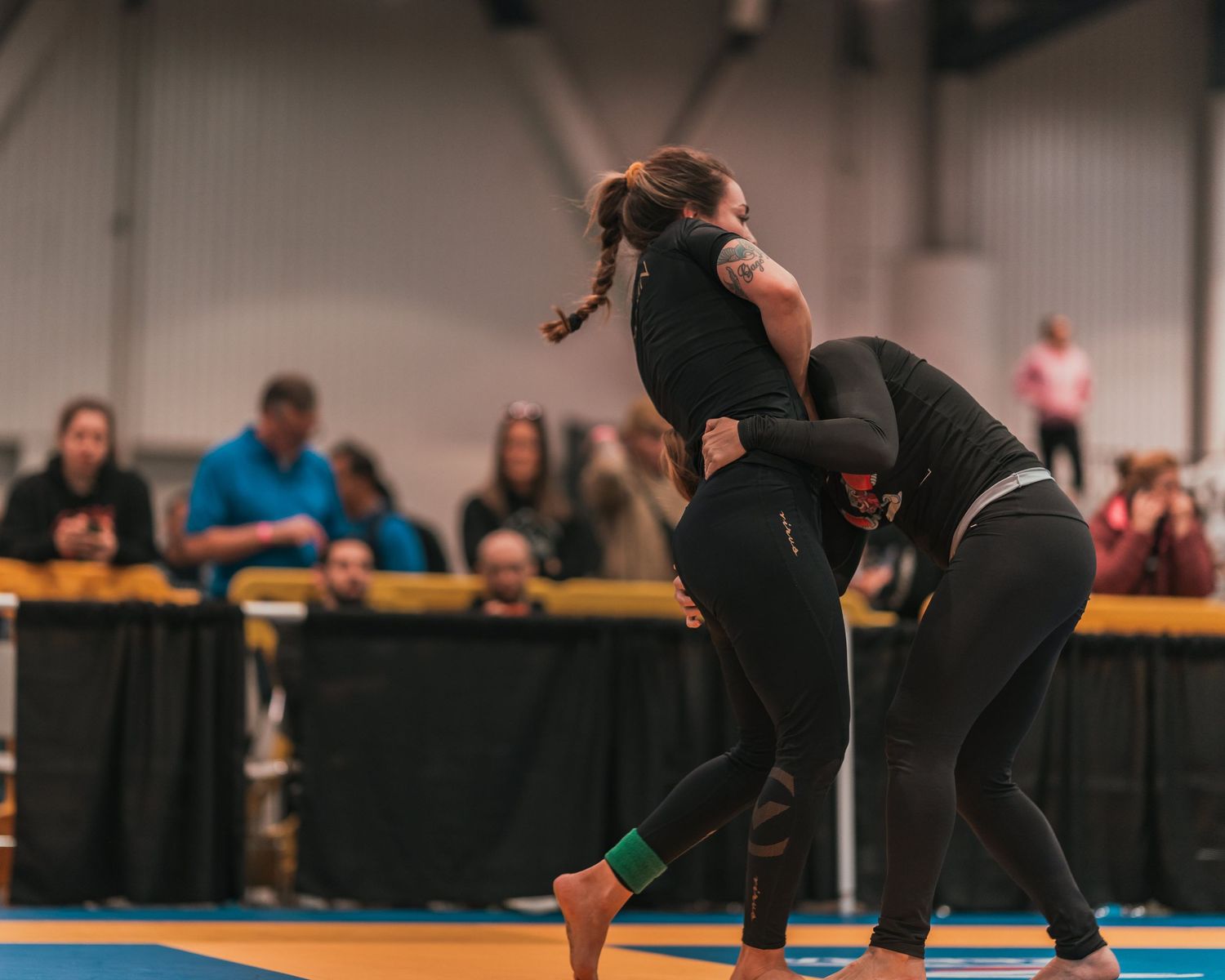 Two competitors grappling during a martial arts tournament with spectators in background.