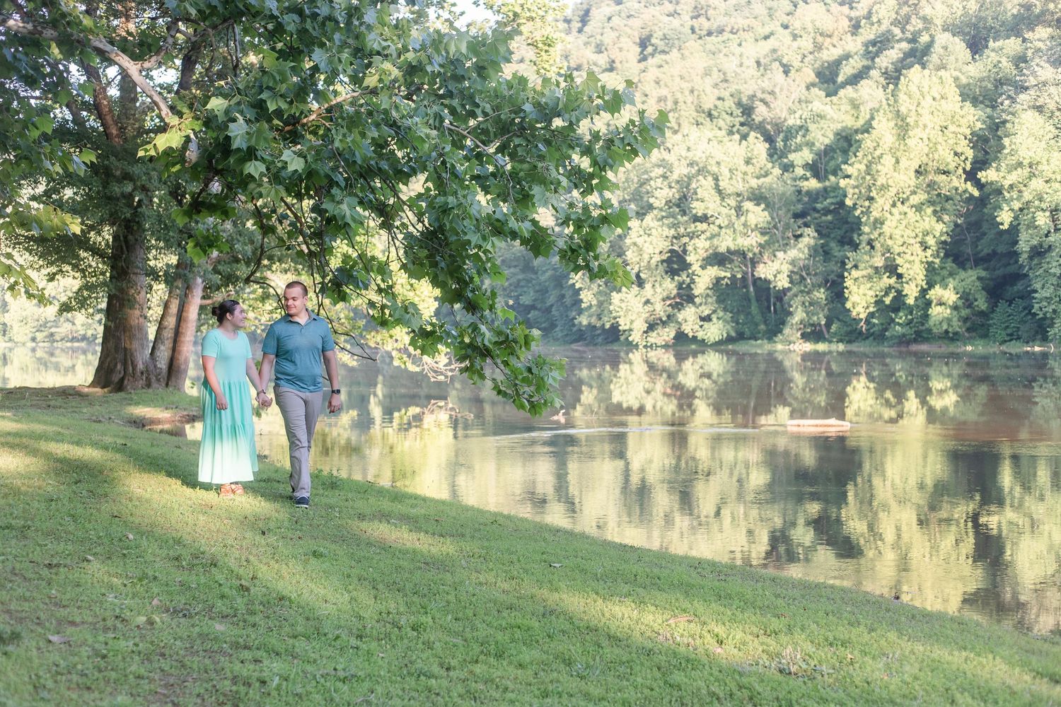 A couple stands together in casual attire by a scenic lake surrounded by lush trees in golden sunlight.