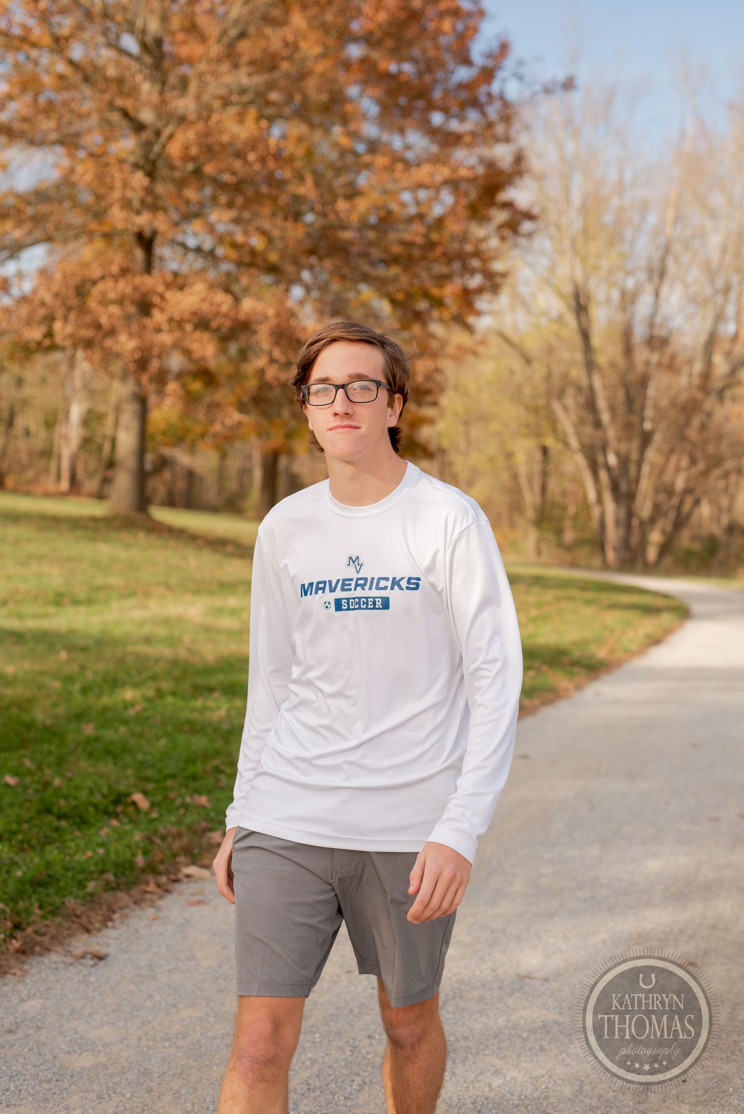 A person in a white Mavericks sweatshirt stands on a path surrounded by autumn foliage.