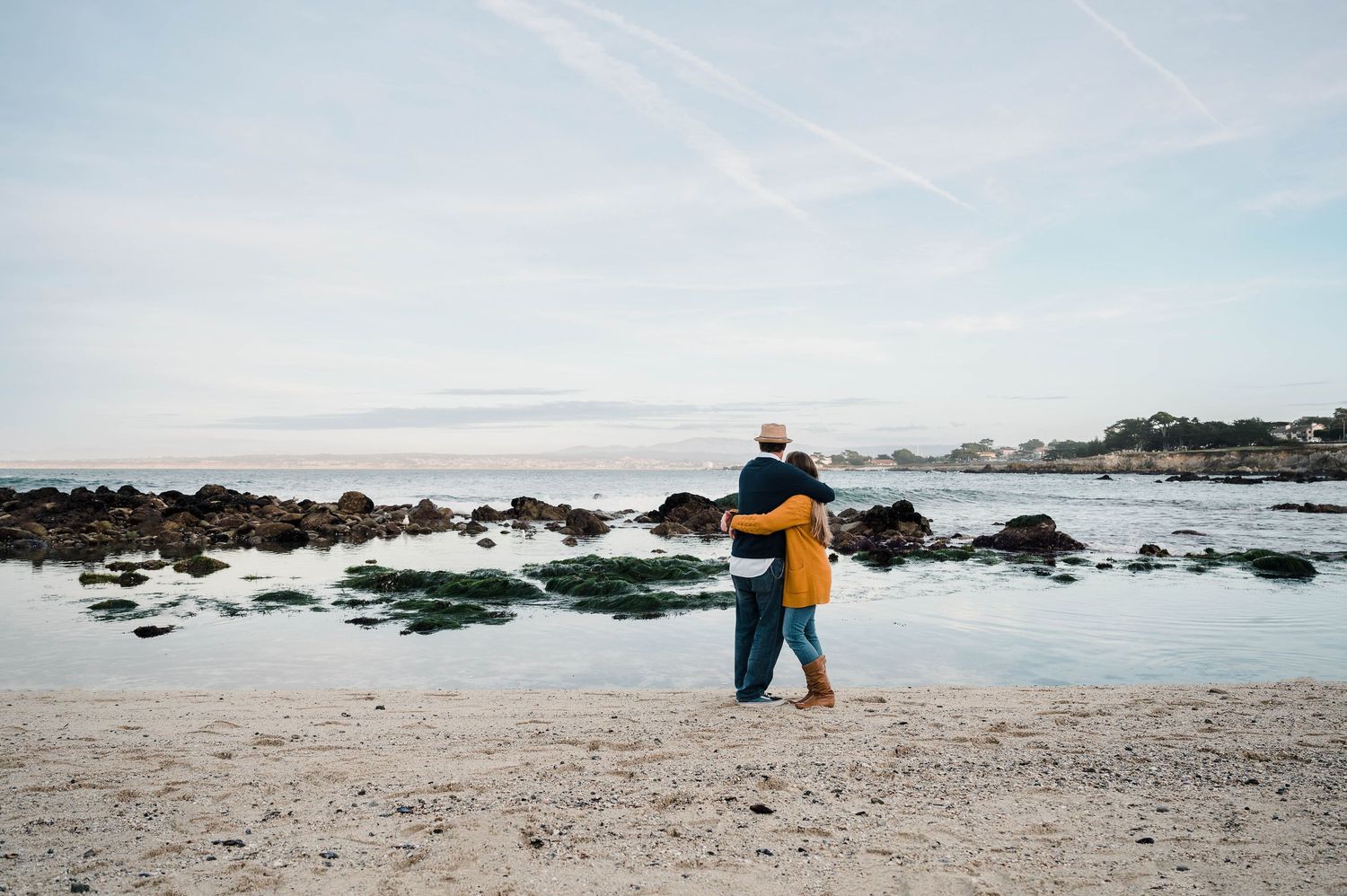 Two people embracing on beach with rocky coastline and ocean view at sunset.