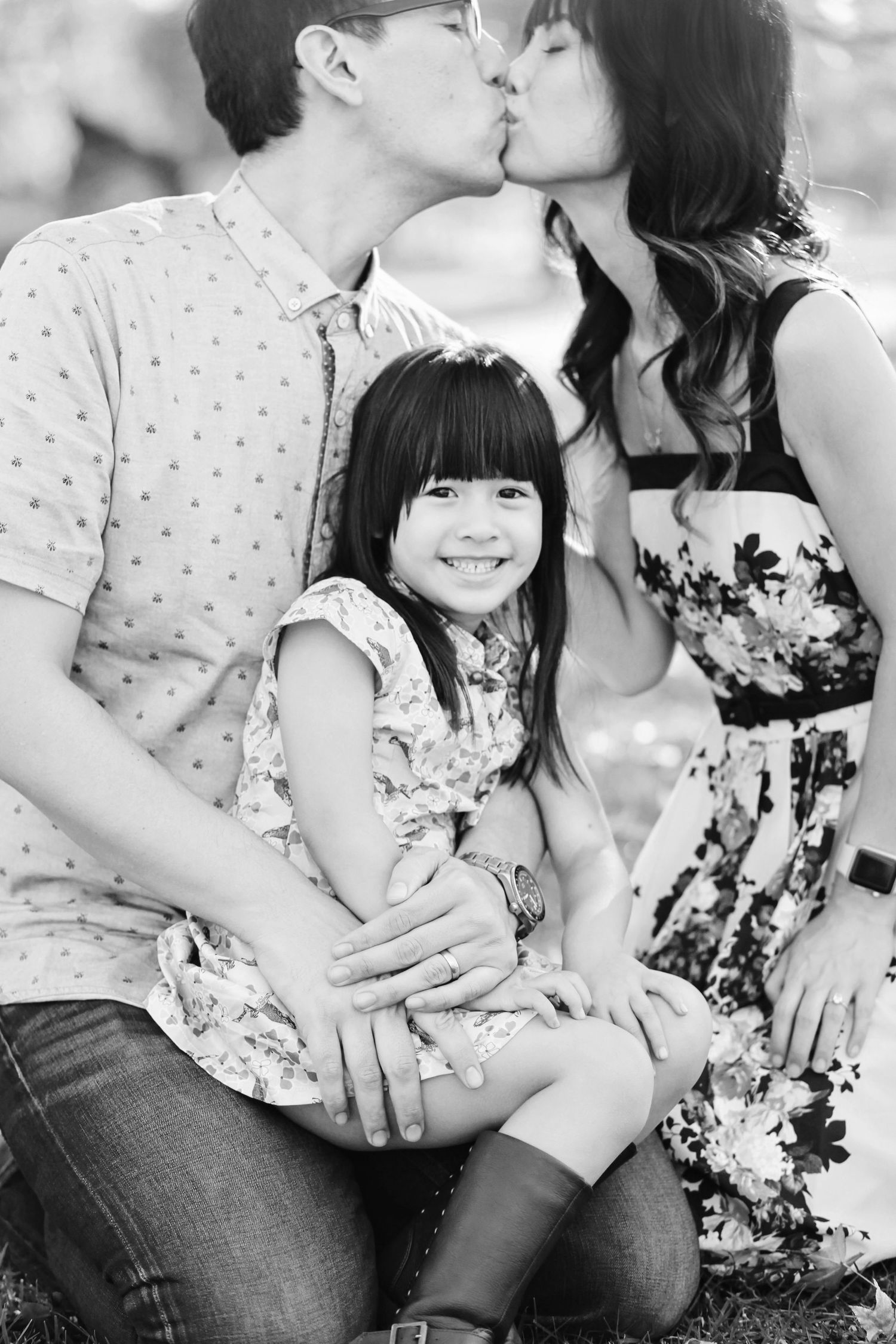 An intimate family portrait with parents sharing a kiss while their young child smiles at the camera.