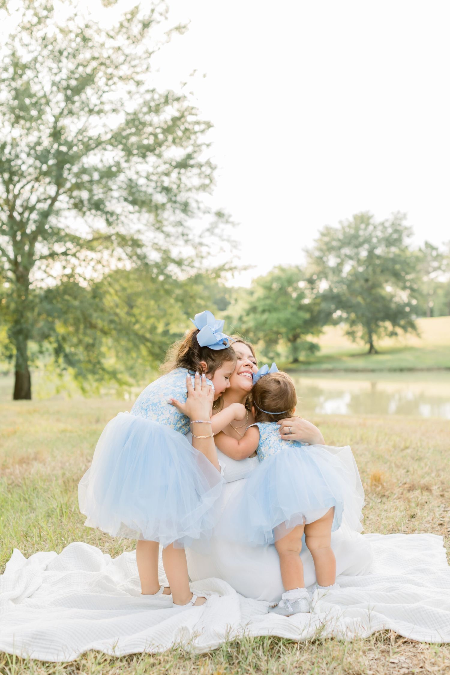 Two small children in matching white tutus hug while sitting on a white blanket outdoors.