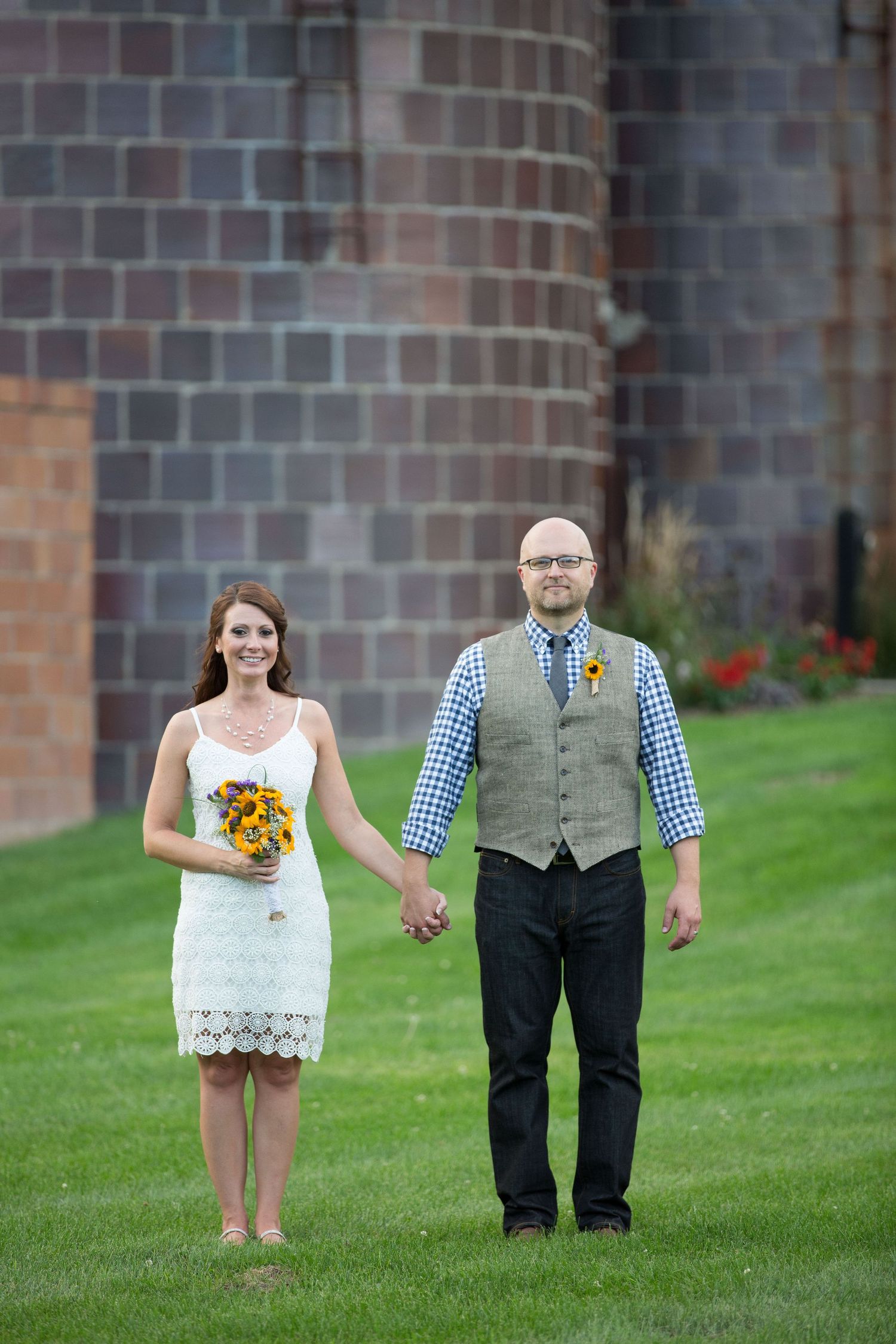 A couple poses on a grassy lawn in front of a brick building with sunflowers in the bride's bouquet.