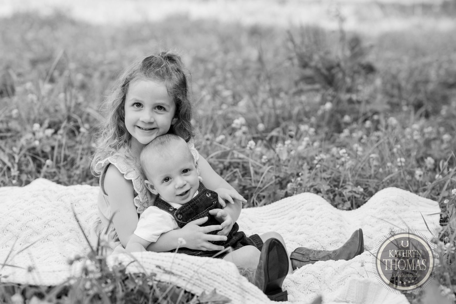 Black and white portrait of two siblings cuddling on a blanket in a wildflower meadow.