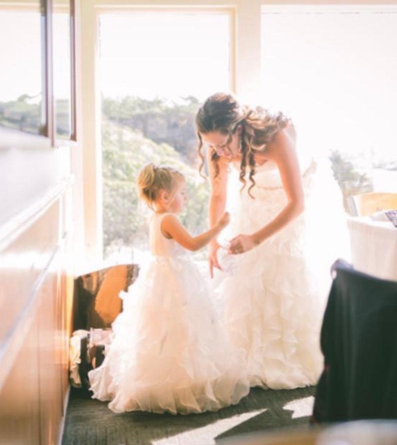 A beautiful bride smiles lovingly at her flower girl, dressed in stunning wedding attire, radiating joy and elegance