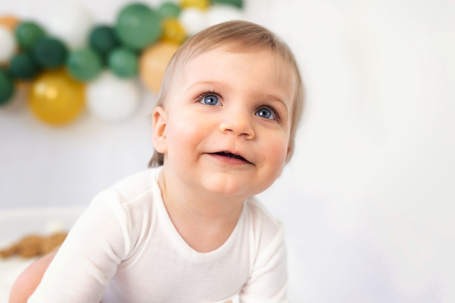 Smiling baby in white outfit sitting against backdrop with green and yellow balloons.