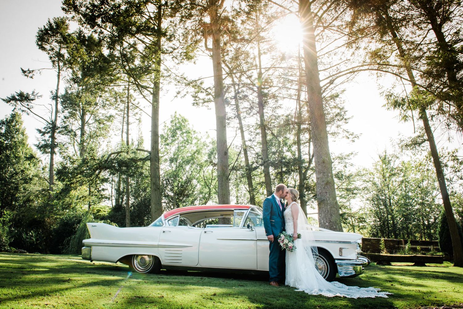 Bride and groom kiss in front of a Cadillac with the sun shining through the trees behind.