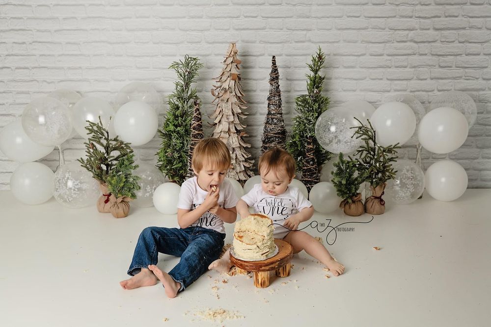 Holiday scene with cake and decorative trees surrounded by white balloons against brick wall backdrop.