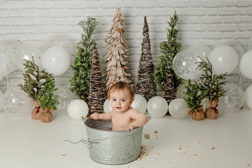 Holiday themed newborn photography setup with rustic decorative trees, white balloons and metallic bucket prop.