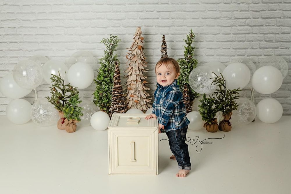 Holiday photo setting with white cube prop, decorative trees and white balloons against brick wall.