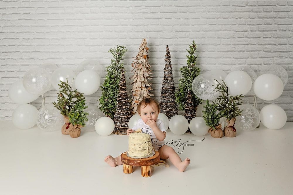 Holiday photo setup with yellow cake, decorative trees and white balloons against white brick wall.