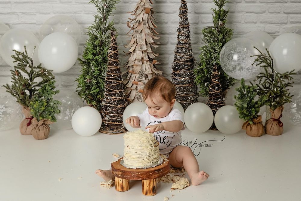 Winter scene with cake on wooden stand, decorative trees and white balloons against white brick backdrop.