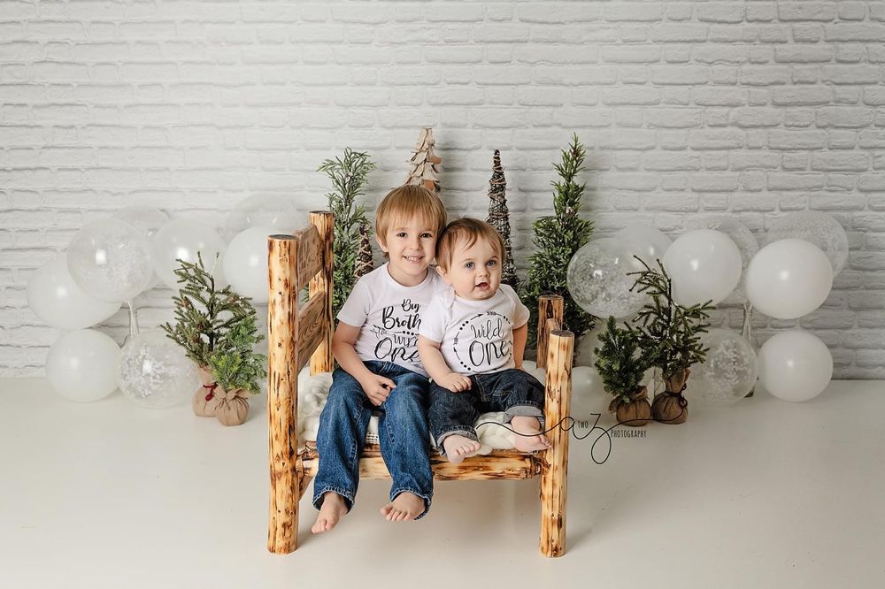 Rustic log bench with decorative trees and white balloons creating cozy winter scene against white wall.
