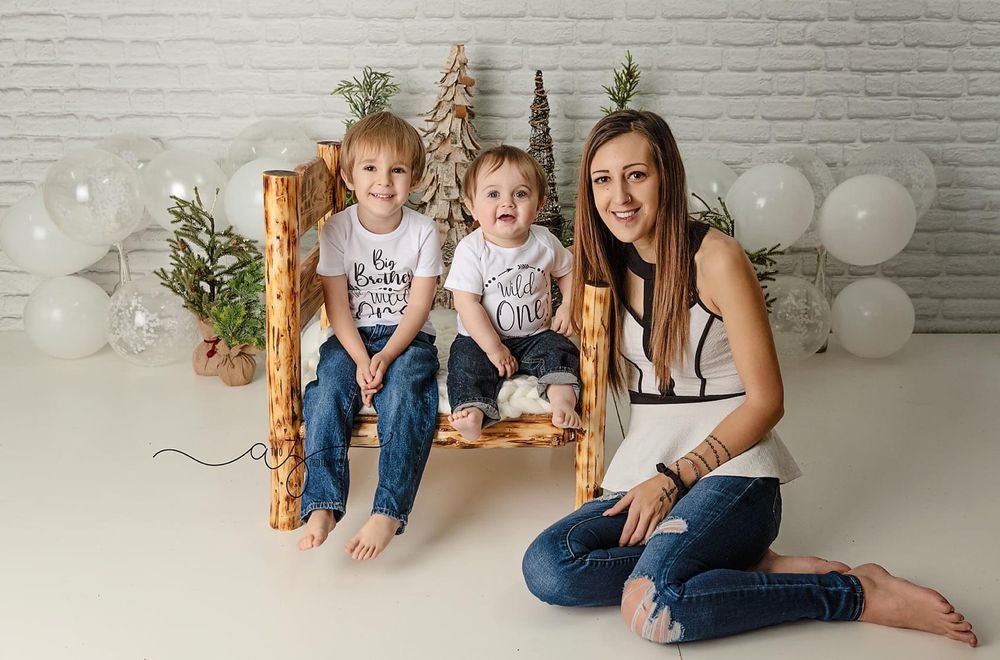 Family winter photo setup with rustic wooden bench, decorative trees and white balloons against brick wall.